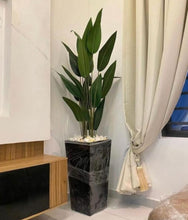Load image into Gallery viewer, Artificial banana plant in a black square planter indoors, with green leaves and a white decorative object at the base.
